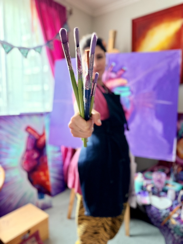 La artista sostiene un manojo de pinceles hacia la cámara, con obras desenfocadas al fondo.